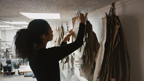 Young multi-ethnic woman, fashion designer or tailor of sewing workshop is choosing a pattern or mold hanging on ceiling. Her colleague working at sewing machine at workstation. Close up