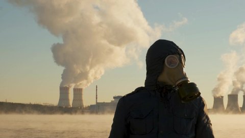 The man in the gas mask looks at the camera, then turns his head to the side. A man stands in front of a nuclear power plant that releases dirty chemicals into the atmosphere. Air emissions, ecology.