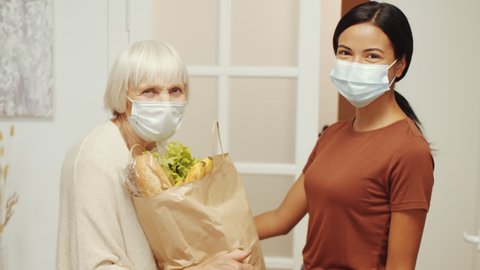 Asian female volunteer in protective mask and gloves giving bag of groceries to senior woman and then posing for camera with her while helping pensioners at home during coronavirus isolation