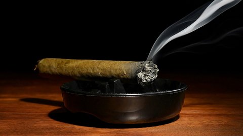 A burning cigar in a classic black ashtray streaming smoking in a dark, moody setting. Shot with a Nikon D7100 24MP camera at f7, 30 shutter speed.