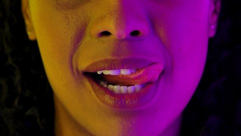 Attractive female African American lips and mouth. Young woman smiles, licks and bites his lips. Close up macro portrait backlit by bright neon lights. Slow motion.