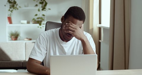 Portrait of afro american student black male tired guy wears white looks at laptop screen learns bad new feels exhaustion stress worry suffers from headache holding his head with hand, problem concept