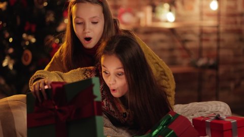 Two exited, Extremely Happy Sisters, Friends opening, unpacking their Christmas Gifts on the Cozy Evening. Two Children in a well-decorated Livingroom near the Christmas Tree looking for Presents.