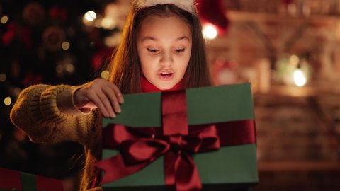 Little Happy Cute Girl is opening her Special Christmas Gift in a decorated room. Caucasian Child celebrating Winter Holidays. Festive Mood. Home Coziness. Christmas. New Year. Happiness.