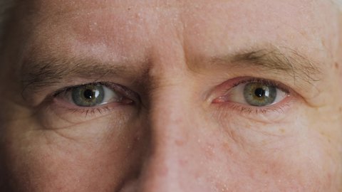 Face of handsome elderly middle aged man opening his blue eyes. Old Caucasian mature male senior citizen with wrinkles on attractive face looking at camera. Slow motion, macro extreme close up, 4K.