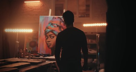 Back view of anonymous man admiring portrait of black woman then walking closer to artwork and applying finishing strokes during work in illuminated workshop