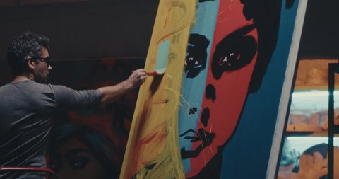 Adult man smearing blue paint over pop art portrait while creating abstract artwork in dark studio