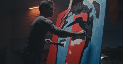 Zoom out view of professional male artist standing on ladder and creating pop art portrait in spacious dark workshop