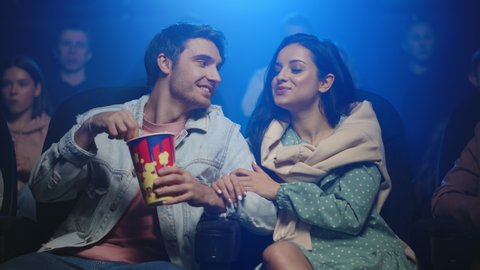Handsome man feeding his girlfriend in cinema. Love couple having fun in movie theater. Smiling woman holding man hand in movie house. Happy people spending date in cinema.