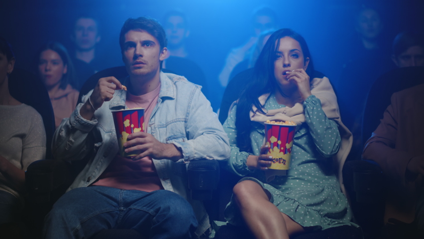 Young people watching film in dark hall. Attractive couple surprising in movie theater. Worried friends sitting with popcorn in cinema. Two friends spending date in cinema. | Shutterstock HD Video #1063803385