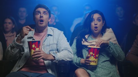 Young people watching film in dark hall. Attractive couple surprising in movie theater. Worried friends sitting with popcorn in cinema. Two friends spending date in cinema.