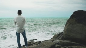 A young brave man in a white shirt and jeans standing on a rock during a storm and looking at the sea