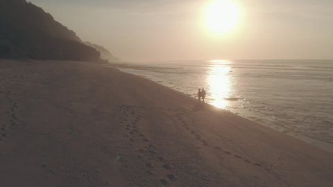 Drone aerial view. Silhouettes of couple in love walking on the beach at sunset. Man and woman holding hands walk, leaving footprints on sand. Sun reflecting in the sea. Concept of romance and love