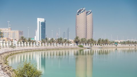 View of skyscrapers skyline with towers in Abu Dhabi timelapse. Reflections on water and palms on the street. United Arab Emirates