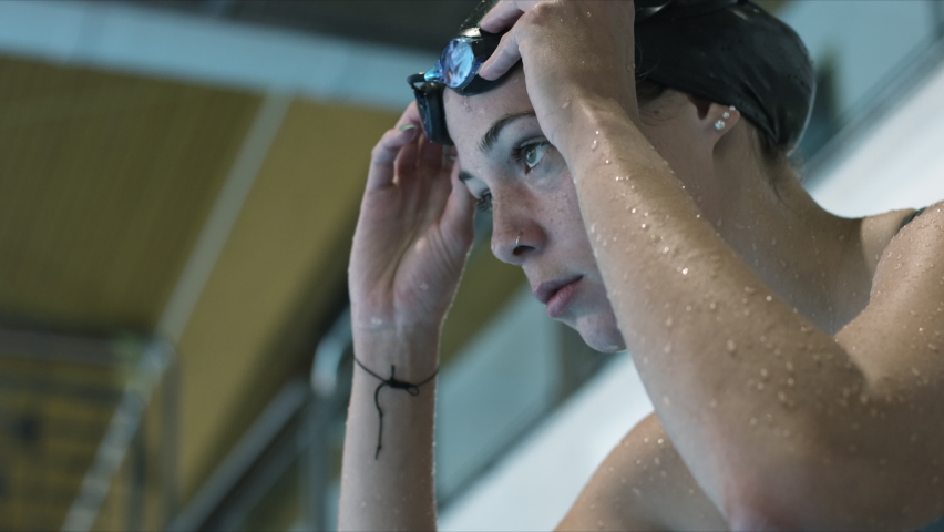 Woman athlete nervously take a deep breath, focus to start swimming, put her goggles on and prepare to jump to the pool. Slow motion video. Top shot. | Shutterstock HD Video #1063818445