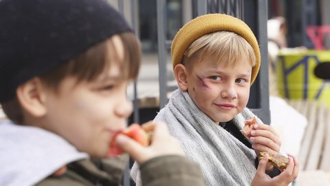 Two homeless boys sit eating food on the bench, little caucasian children, street kids are hungry, get some food by kind people.