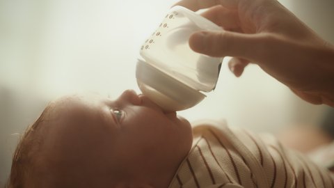 Authentic Close Up Footage of a Newborn Drinking Milk from Small Baby Bottle while Lying on the Back in Child Crib. Portrait of Caucasian Neonate Toddler at Home. Concept of Childhood and Parenthood.