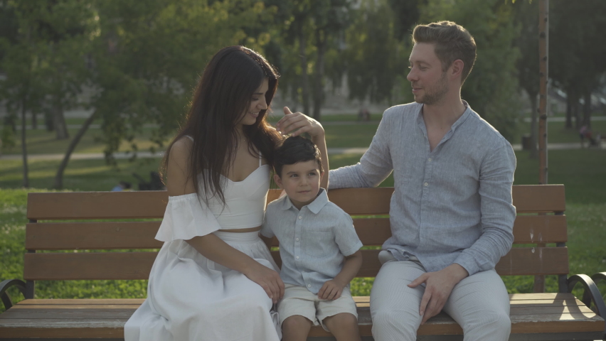 Happy interracial family sitting on bench in sunny park and smiling at camera. Portrait of happy young Caucasian man, beautiful Middle Eastern woman, and cute mixed-race boy posing outdoors.