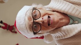 Vertical video of Elderly woman with Santa hat, looking at camera, talking fast and happy on video chat on conference call. Wishing merry Christmas to their relatives. Self isolation quarantine. POV.