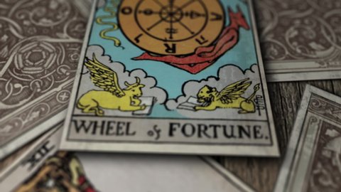 Tarot cards falling on a wooden table. One of the cards is revealed and it says wheel of fortune, with a compass in the middle and different figures of creatures with wings. Mystic game. Close up