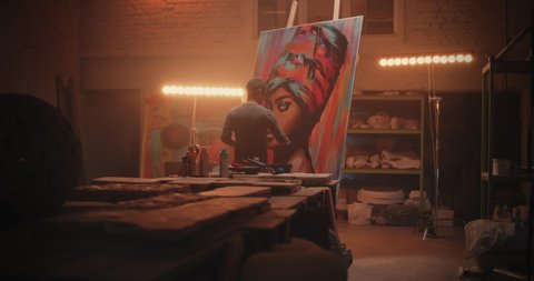 Zoom in view of male artist smearing paint on portrait of African woman while working in illuminated workshop