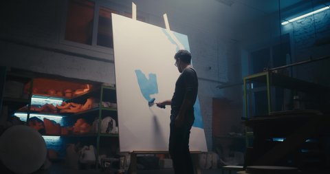 Zoom in of creative man smearing blue paint with roller on paper sheet while working in dark professional studio