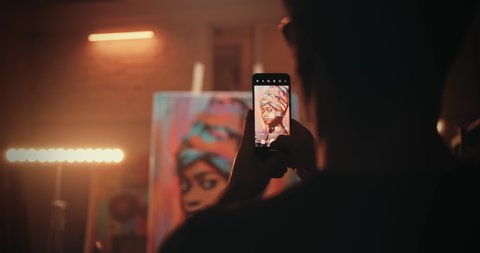 Rack focus of crop painter taking photo of colorful portrait of African woman behind workbench in illuminated workshop