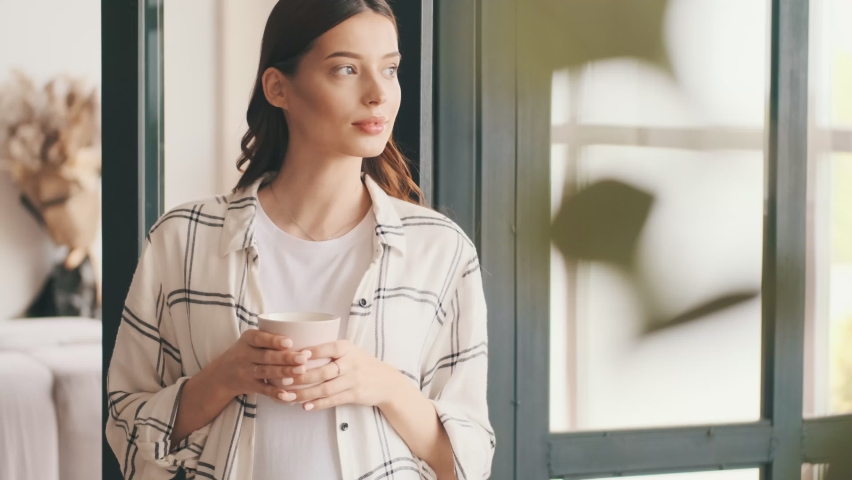 Positive pregnant woman looking at the window while drinking coffee indoors at home | Shutterstock HD Video #1063831021