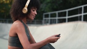 Young african sports woman using mobile phone outdoors in park while listening music in headphones