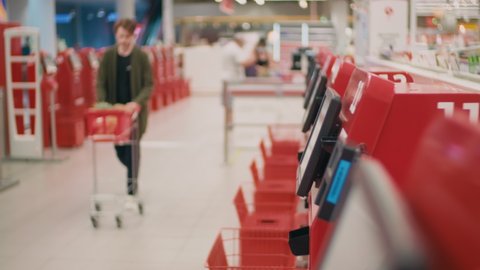 Medium shot of young man with cart purchasing food products using self-checkout system scanning items in hypermarket