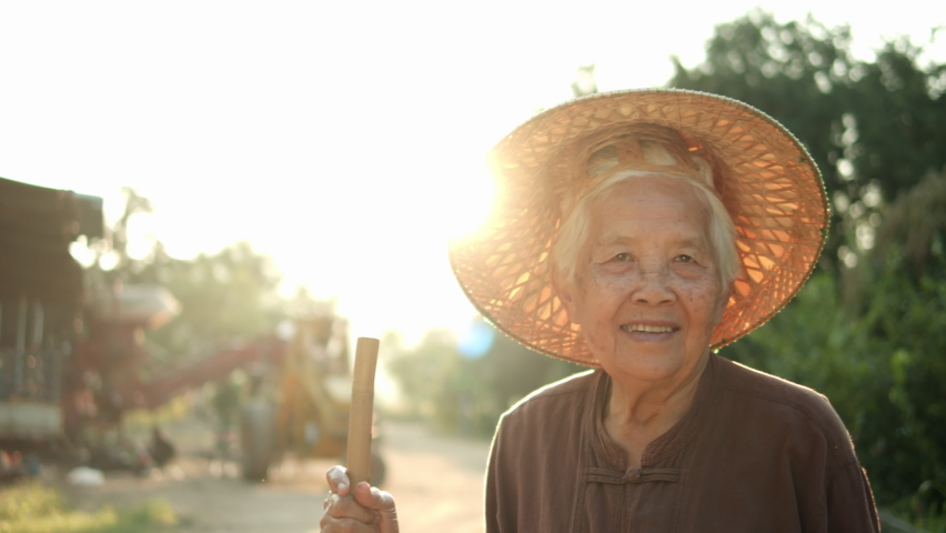 Slow motion of the smiling elderly Asian female farmer has bright white hair, wear weaving hat and local woven garment ,standing with handle of a shovel in the evening, the sun shone from behind her. Royalty-Free Stock Footage #1063837837