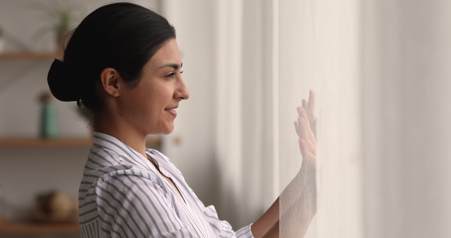 Close up side profile view Indian attractive woman opens curtains in own apartment in morning. Young pretty female enjoy weekend alone, city view through window feels carefree. New happy day concept Royalty-Free Stock Footage #1063839247