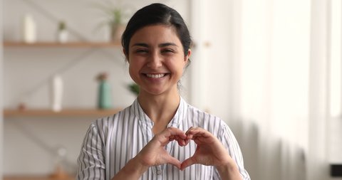 Headshot portrait 25s Indian woman show heart sign make symbol with hands feels grateful smile looks at camera standing indoors. Love support demonstration, volunteering, donation, good deed concept