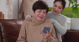 Young beautiful Asian girl sitting on the sofa giving her mother shoulder massage Happy middle-aged elderly woman holding mobile phone shopping online with adult daughter family lifestyle 4k clip