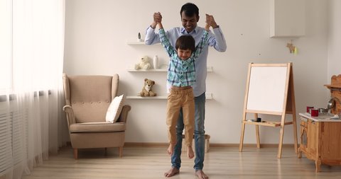 Little 5s Indian preschooler boy jumping holding loving father hands having fun in warm modern cozy living room. Family enjoy carefree weekend together, happy fatherhood, wellbeing, playtime concept