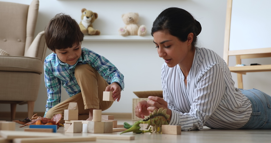 Indian nanny spend time in modern cozy home with little boy. Loving caring mother play with preschooler son with dinosaurs, construct tower use wooden bricks set. Child development, playtime concept Royalty-Free Stock Footage #1063839394
