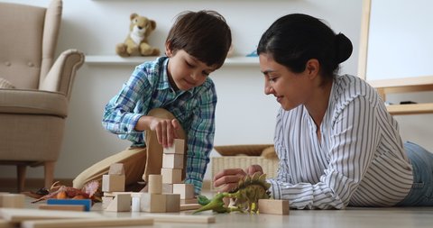 Indian nanny spend time in modern cozy home with little boy. Loving caring mother play with preschooler son with dinosaurs, construct tower use wooden bricks set. Child development, playtime concept