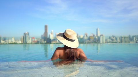 Backside view of a Pretty young model wearing a swimming suit and hat and sitting inside infinity pool enjoying the city view, hotels rooftop