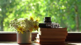 Closeup view 4k video of windowsill of rural cottage. Bouquet of cute small field flowers in blue metal mug, several paper books and old photo camera laying on window sill.