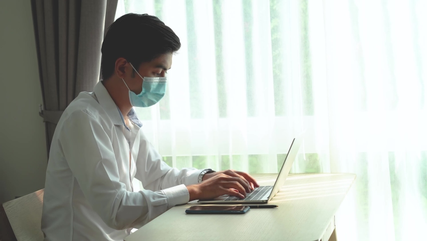 Young asian man in protective facial mask taking a call on his mobile phone checking information received on his laptop with a serious concerned expression | Shutterstock HD Video #1063844332