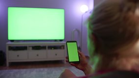 blonde woman sitting on sofa bed doing scrolling on smart phone green screen big screen television in the background