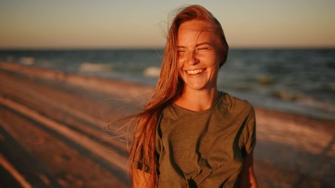 close up face portrait happy woman in khaki t-shirt hair fluttering blow wind. young thin beautiful blonde woman laughs smiling rejoices having fun radiating health sea on beach sunset in front camera