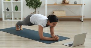 Indian young slim woman watch video on laptop perform butt workout plank leg raises on mat. Fitness for weight loss, burn gluteus muscles, improve shape. Sport from home using on-line training concept