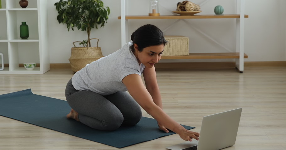Pretty Indian woman wear activewear sit on mat in lotus position turns on laptop relaxing music for stress relief do meditation practice. Sport at home due corona virus, improve mental health concept Royalty-Free Stock Footage #1063846972