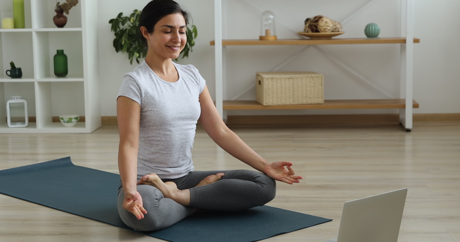 Pretty Indian woman wear activewear sit on mat in lotus position turns on laptop relaxing music for stress relief do meditation practice. Sport at home due corona virus, improve mental health concept | Shutterstock HD Video #1063846972