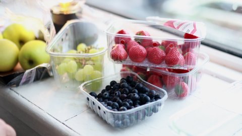Female hands pouring blueberries and strawberries into plastic container above kitchen counter. Woman holding fruit in plastic box. Fruity dessert preparation