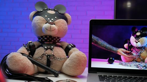 toy bear in a leather belt accessory for BDSM games next to a laptop TV in neon colors