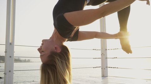 Flexible blonde woman in modern tracksuit turns and does exercises hanging upside down on aero yoga sling on pier ground near tranquil sea at sunset