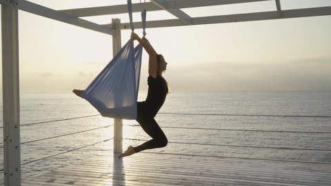 Athletic blonde woman in comfortable tracksuit does yoga asana hanging on aero yoga sling on pier ground near tranquil sea water at sunset light