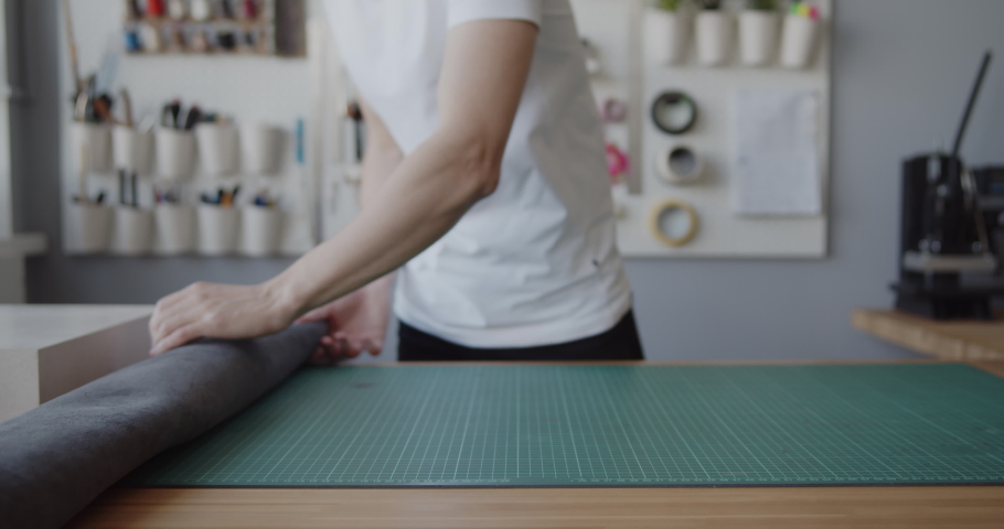 Young leather worker examines a large black leather piece on a working desk in his workshop. Runs his hand over the surface checking its quality.